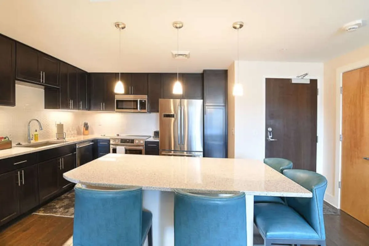 A bright, modern kitchen featuring a spacious island with bar stools, gleaming stainless steel appliances, stylish pendant lights, and ample counter space. The setup allows for both cooking and socializing, with areas clearly suited for entertaining guests or enjoying casual family meals.