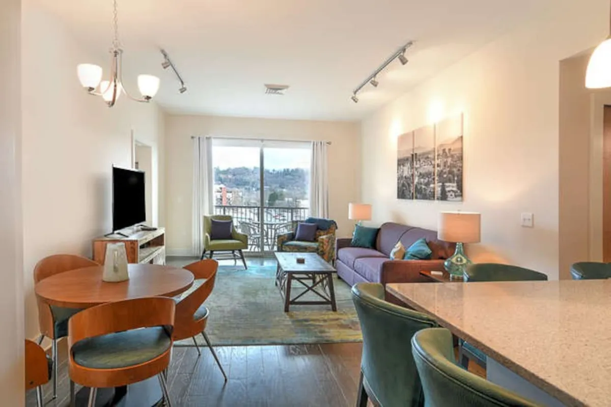This image showcases a spacious, sun-soaked open-plan living and dining area. The focal points include a comfortable sofa—likely serving as the queen sleeper—positioned near a coffee table, with a dining table and chairs set nearby. Large windows bathe the space in natural light, and a TV and balcony view contribute to the inviting, communal atmosphere. The setting is arranged for both relaxation and socializing, perfectly aligning with the section's emphasis on gatherings.