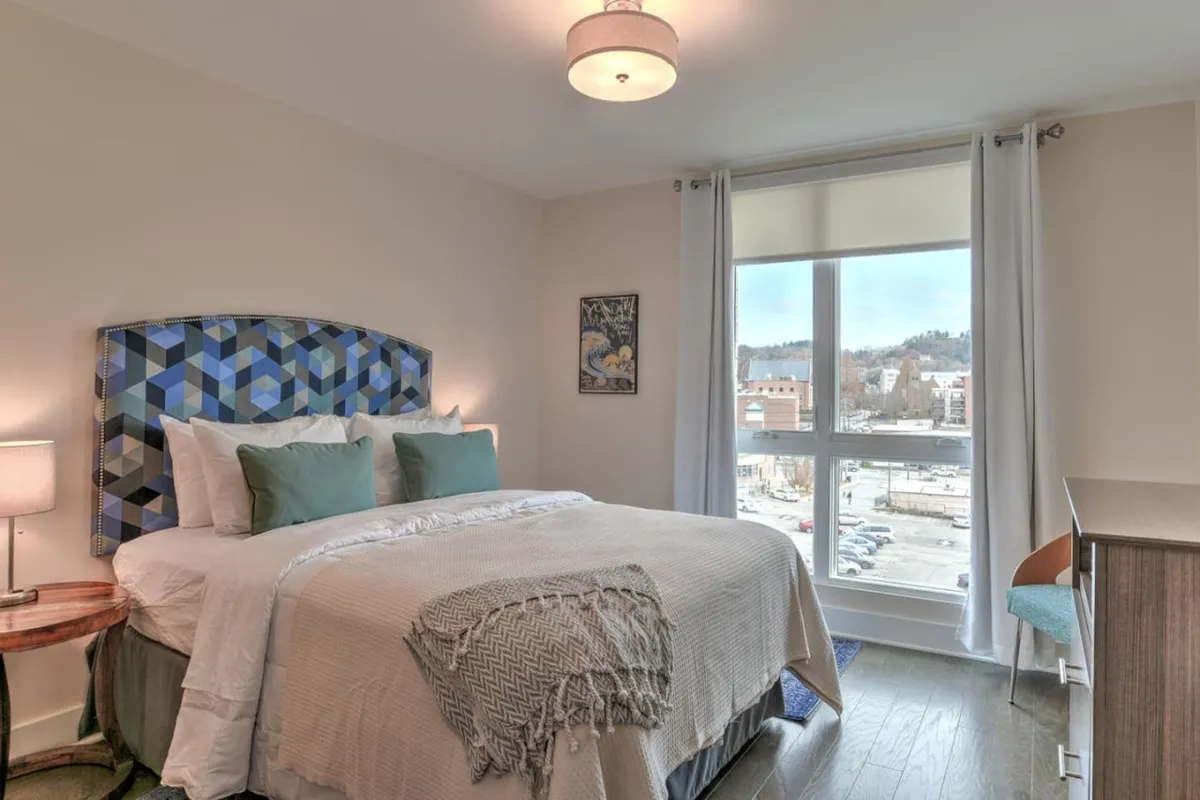 A cozy bedroom featuring a modern geometric headboard, a large window that offers a view of the city, contemporary wall art, and a warmly-lit nightstand. The inviting atmosphere suggests both comfort and a connection to the vibrant world outside.