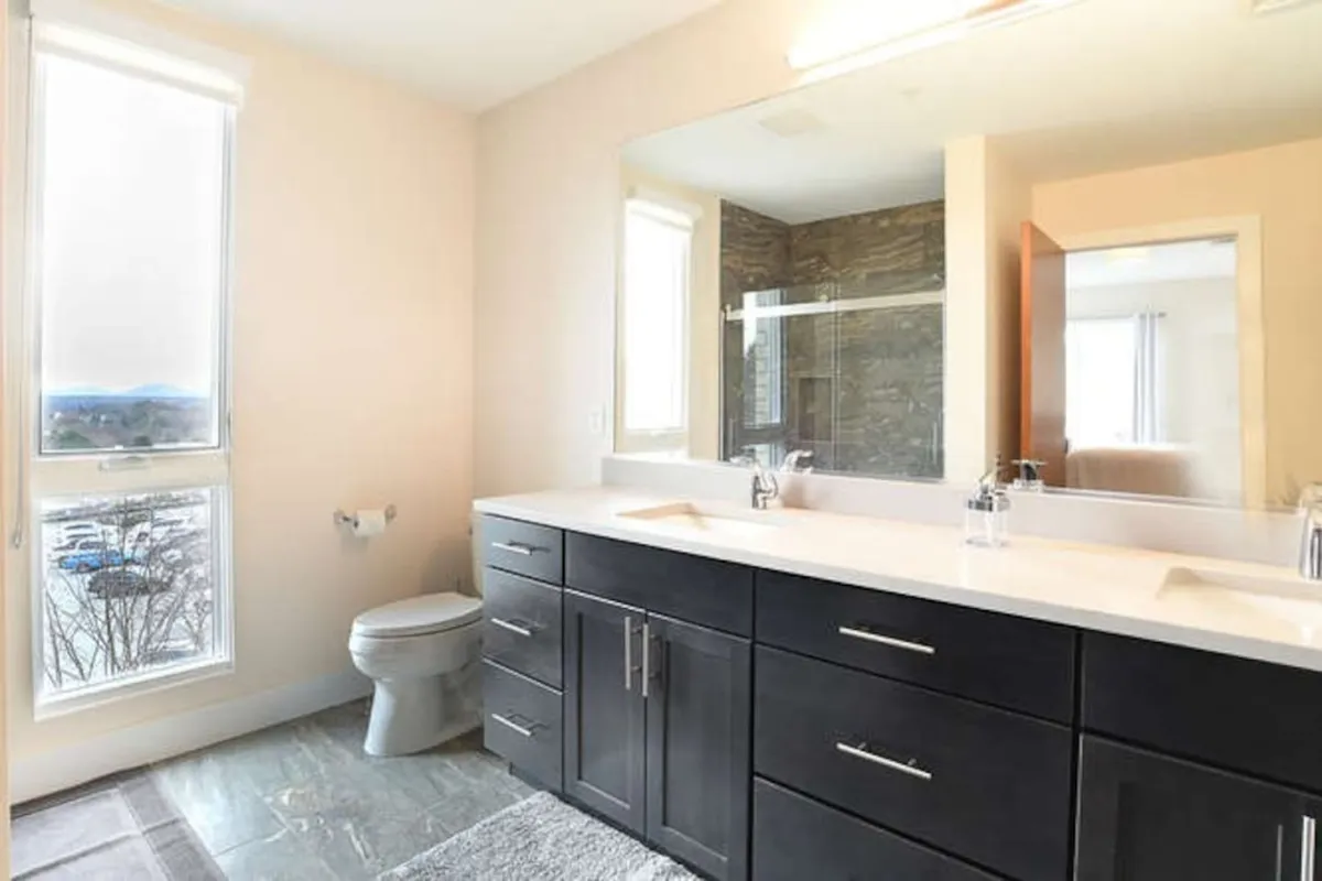 A spacious, modern bathroom featuring a double sink vanity, expansive mirror, shower enclosure, and a window offering daylight. The space feels refreshing and well-appointed for morning or evening routines.