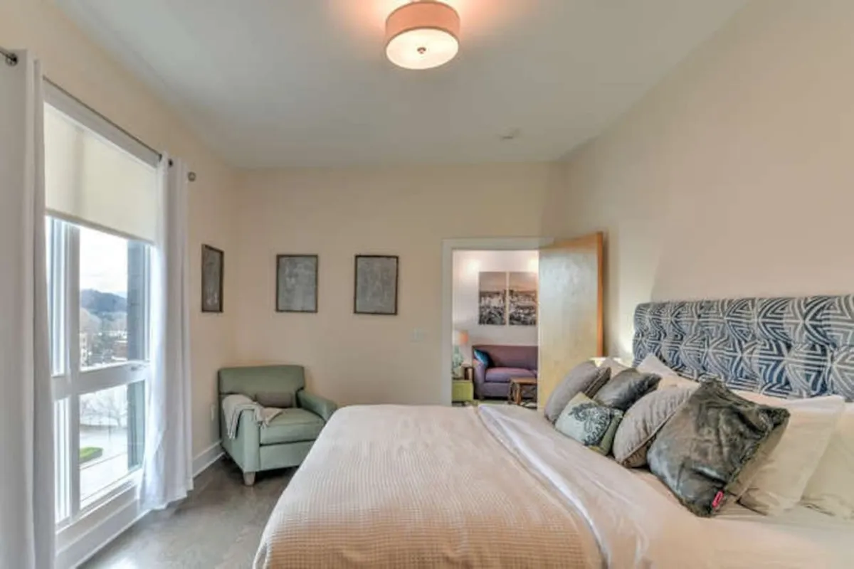 A serene bedroom featuring a plush bed with decorative pillows, a cozy green armchair in the corner, and framed artwork adorning the walls. Soft natural light streams in, creating a tranquil space ideal for relaxation or enjoying a morning coffee.