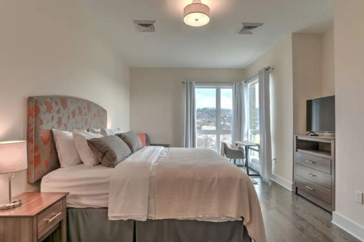A bright queen bedroom with a decorative headboard, large window letting in natural light, and a modern TV unit. The setting exudes serenity, with a bed perfect for cozy evenings or leisurely mornings.
