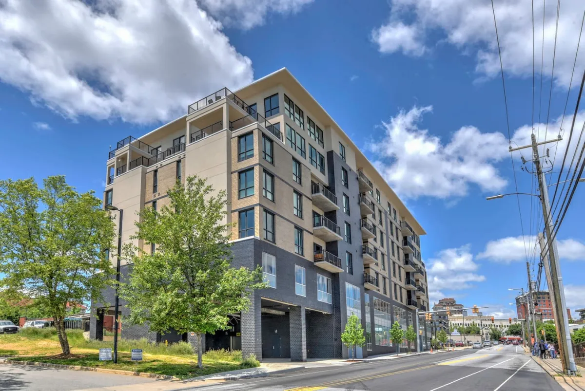 This image captures the building’s contemporary exterior, featuring striking modern architecture with large windows and lush green trees framing the entrance. The scene hints at community activity with visible pathways for walking or cycling, and the vibrant neighborhood atmosphere is further suggested by the warm light and inviting facade.