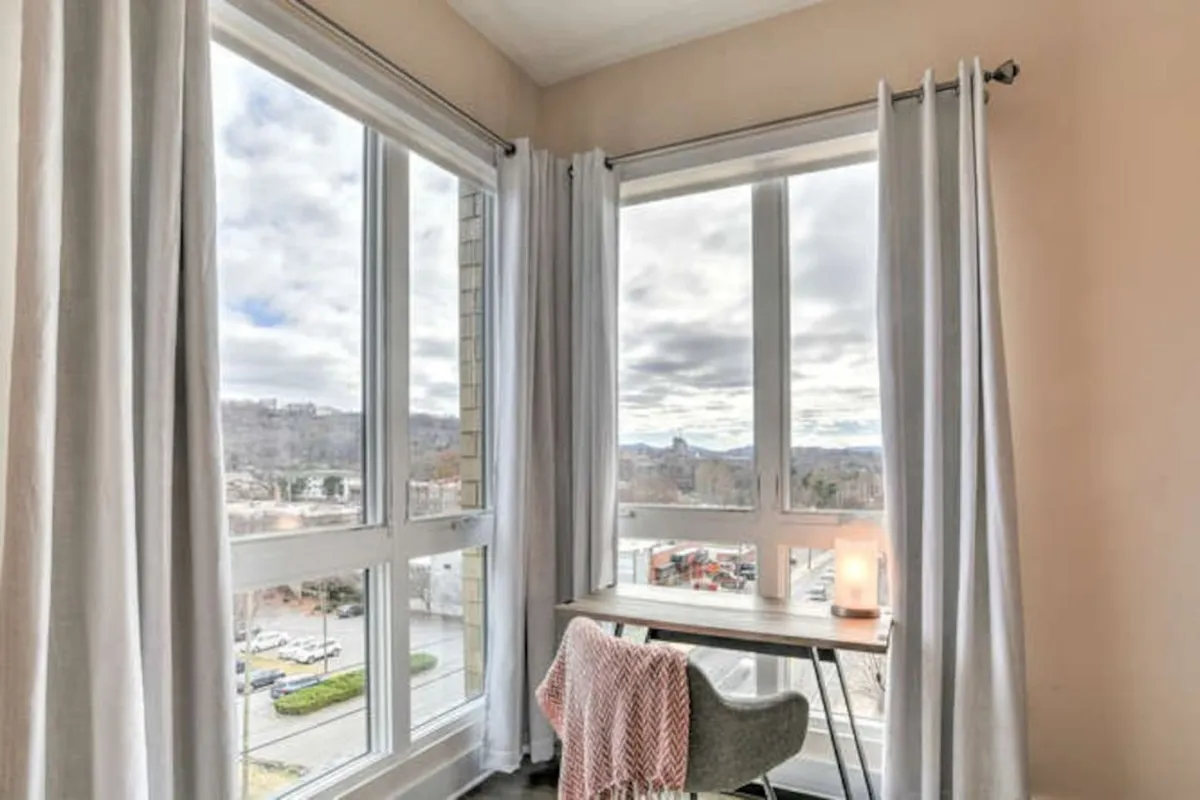 A modern, light-filled work nook featuring a large window, a sleek desk with a lamp, and curtains for privacy. The space is designed for productivity but maintains a calm, inviting mood that flows with the adjoining living and dining area. The setup encourages remote work or trip planning without feeling isolated from the communal environment.