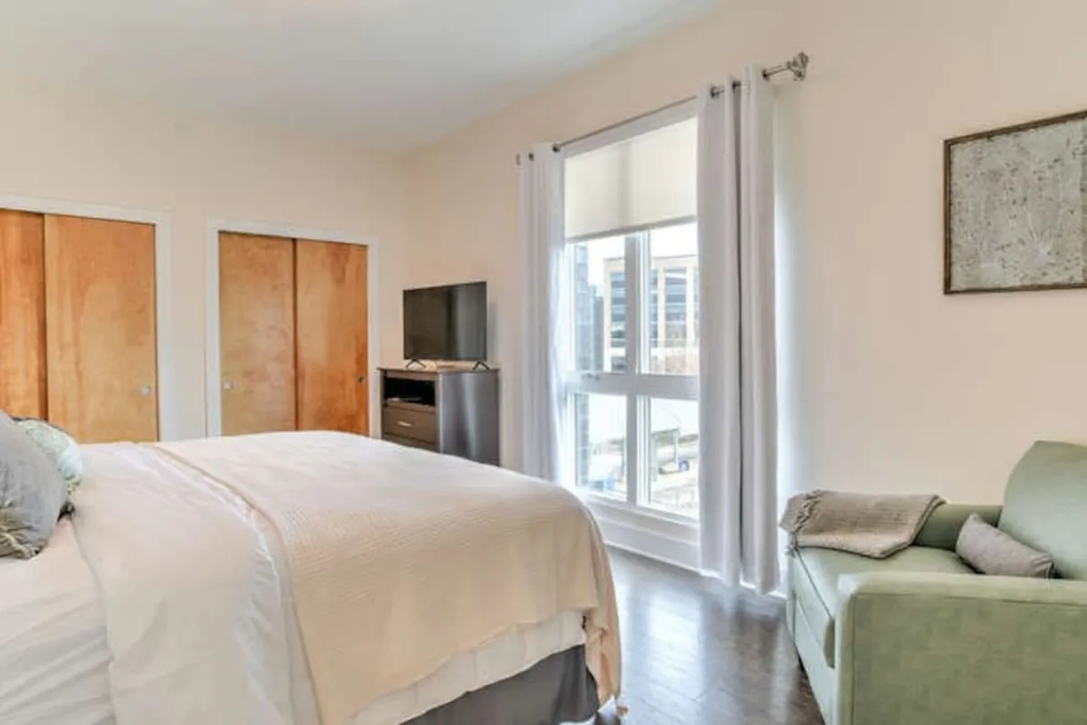 A peaceful bedroom scene with sunlight streaming through a window, a comfortable bed, and a cozy chair perfect for unwinding. The window hints at the vibrant life outside, aligning with the ease of access to local attractions.